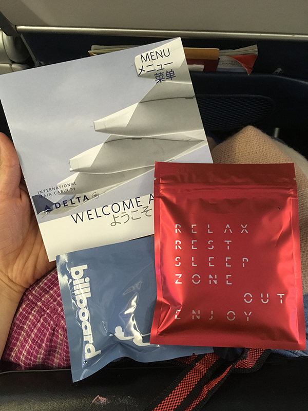 Image of Delta's economy seat amenities: a menu, eyemask, earplugs, and earbud headphones (not pictured: blanket, pillow, and slippers)